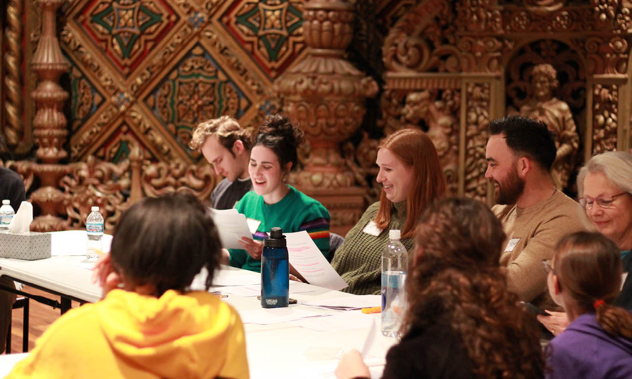 Group of young people sitting around a set up tables reading scripts aloud and interacting