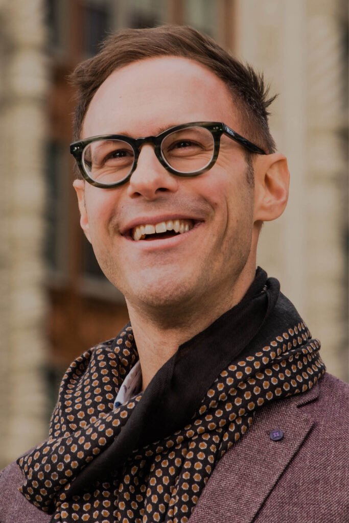 Headshot of a caucasian man with glasses and a scarf smiling and standing in a city scape.