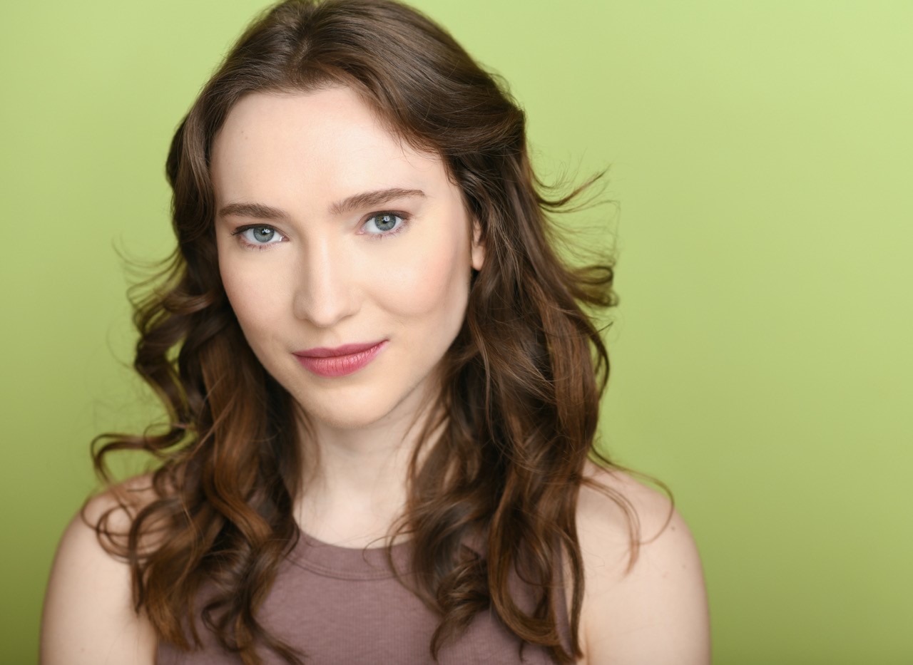 Portrait of a young, Caucasian woman with long brown hair wearing a brown shirt in front of a green background