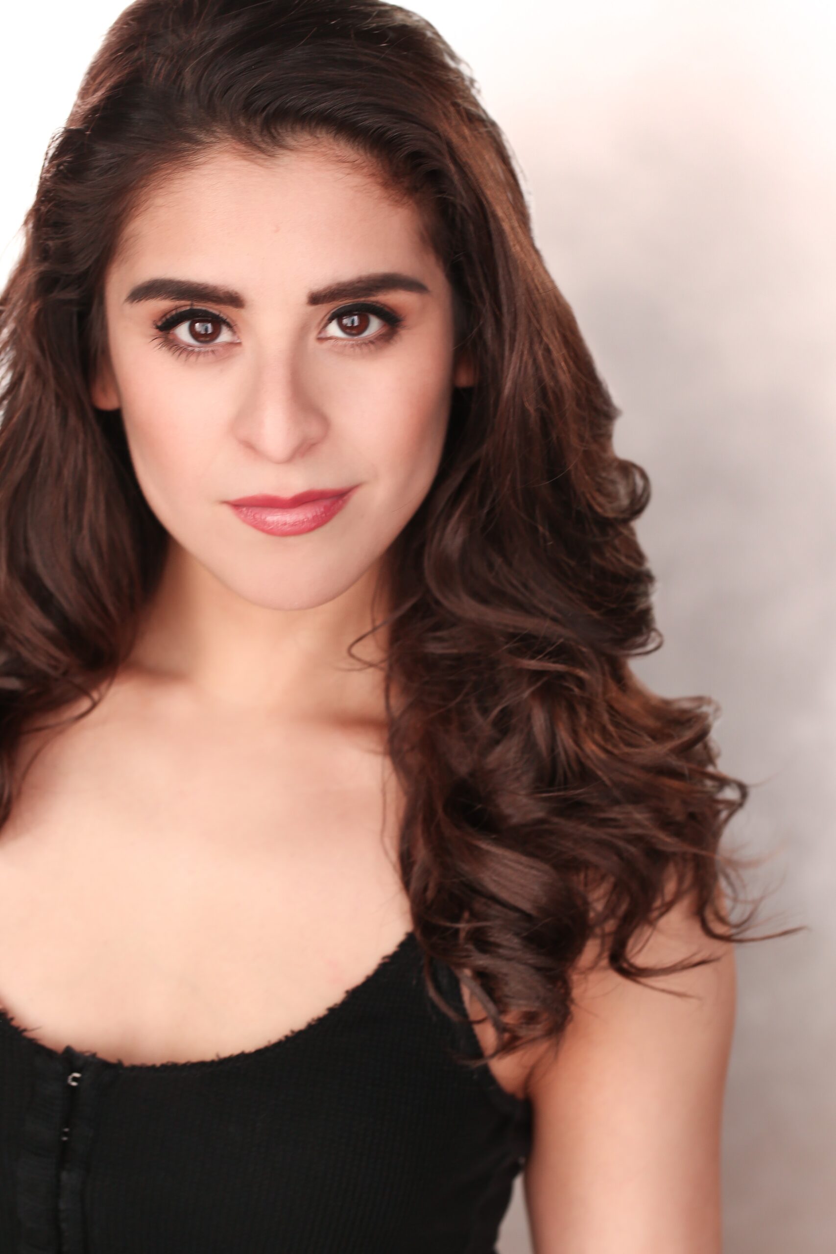 Headshot of a young, Hispanic woman with long brown hair wearing a black shirt in front of a light-colored background