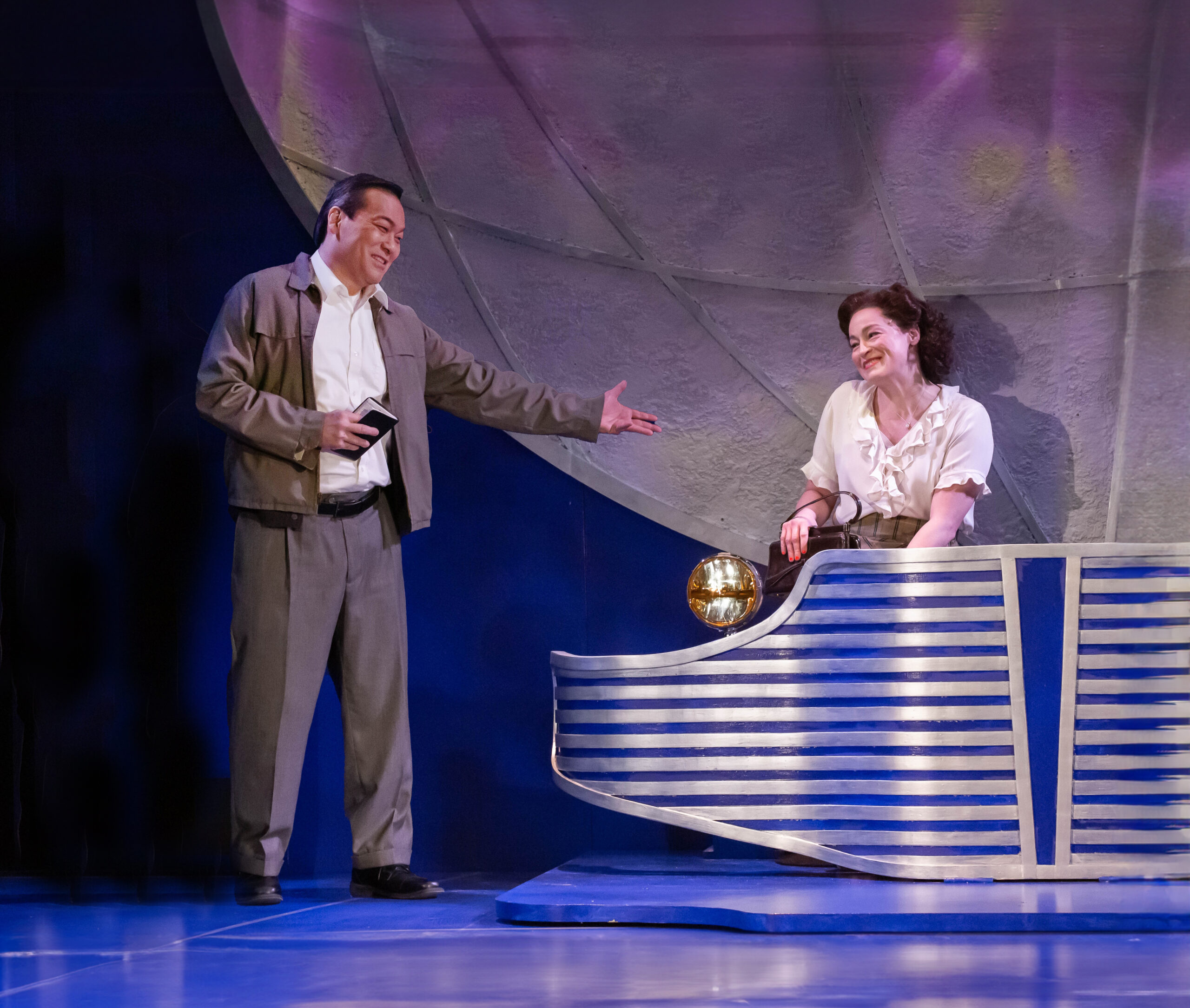 Photo of person sitting in a model of a car, with another person gesturing to the passenger.