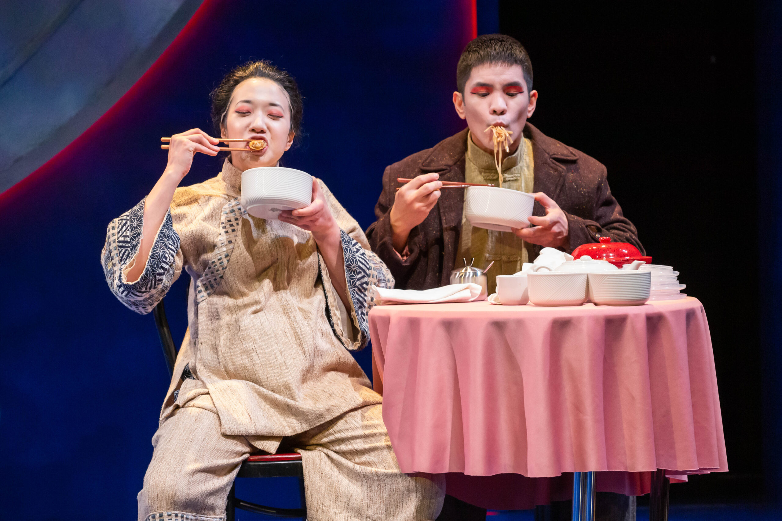 Photo of two people sitting at a table with a pink cloth, eating Chinese food with chopsticks.