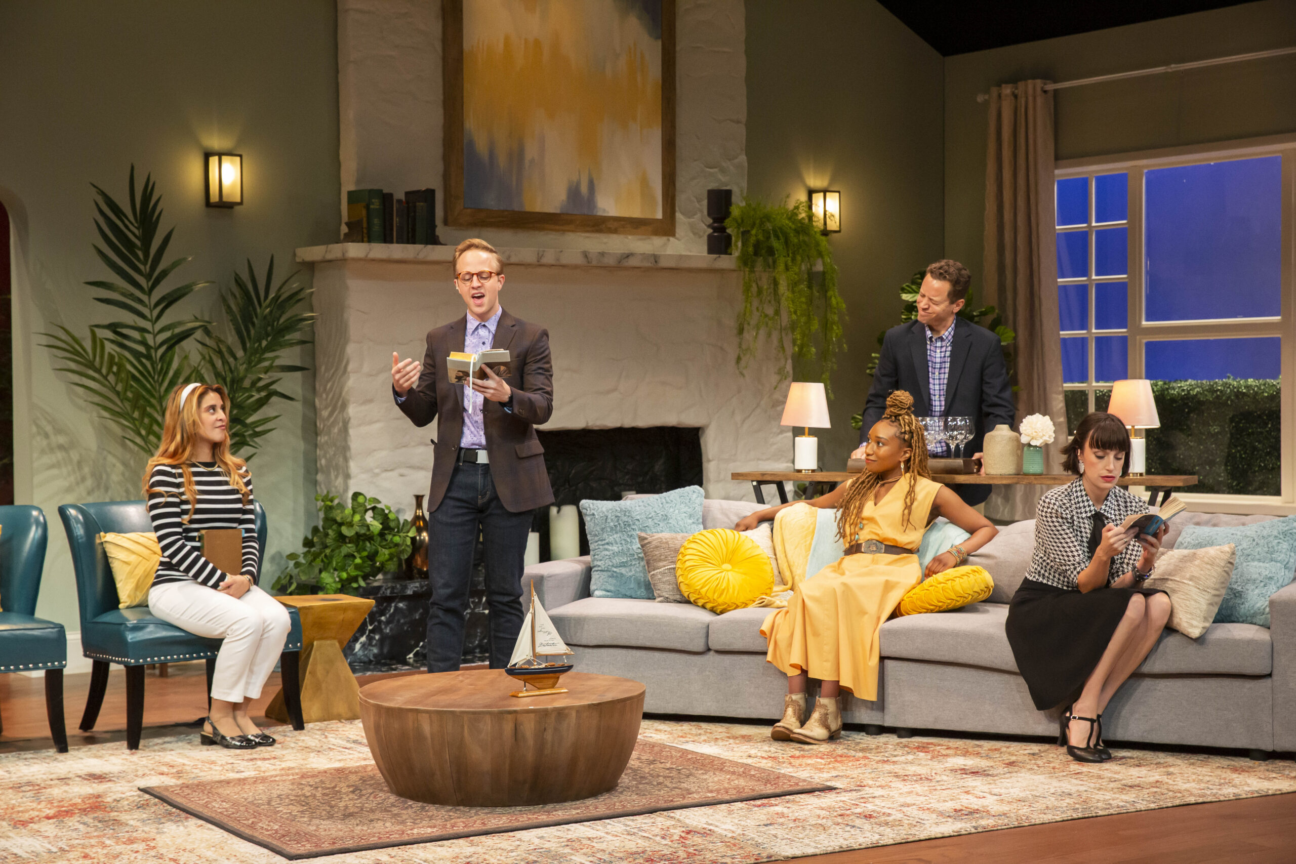 Photo of a group of five people sitting together in a modern living room, listening to one person read aloud.